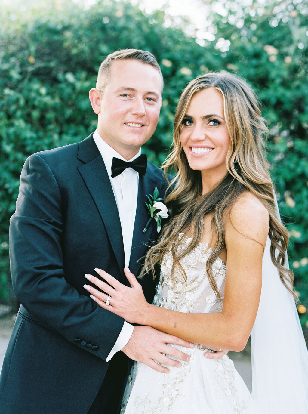 The bride and groom pose for an elegant wedding portrait against lush greenery, capturing the essence of timeless wedding photography with natural beauty and classic style.