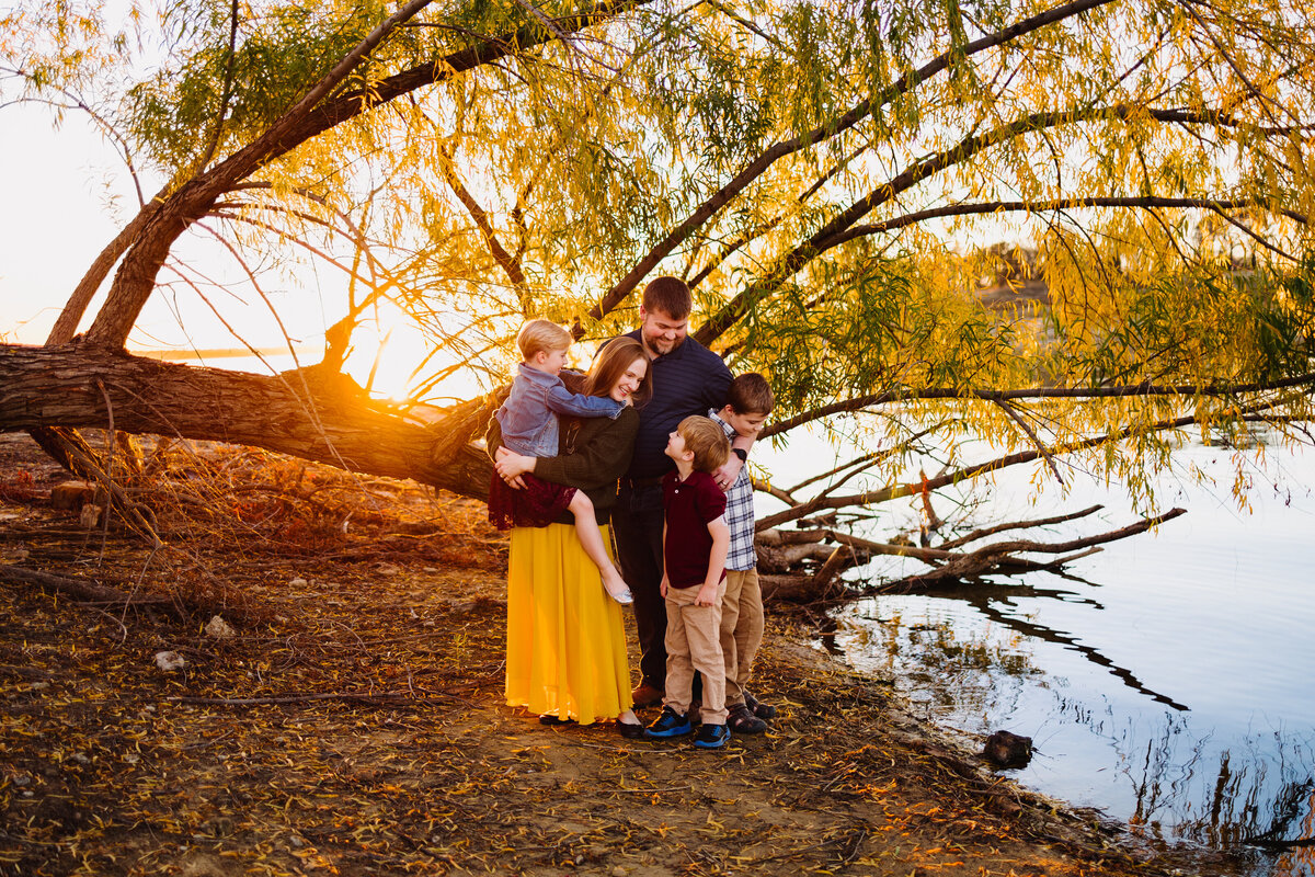 Family photo on a rock, behind them is a tree with many green leaves. The yellow skirt woman is carrying a child who is looking down to the floor while the man sees the other two boys dressed in blue shirt. In front of them is the edge of a lake