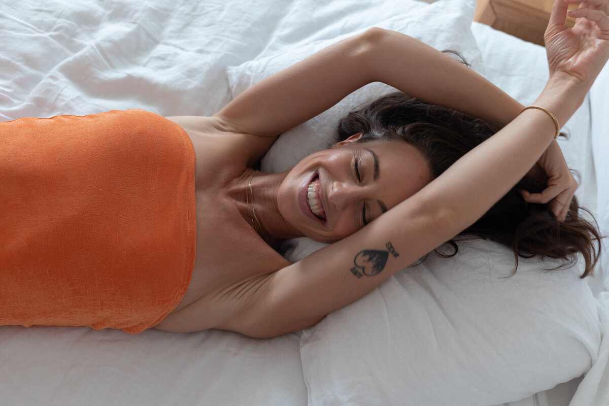 Meg O'Neill laying on the bed wearing orange dress smiling with her hands in the air