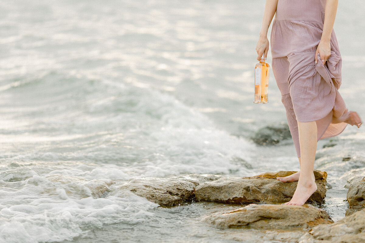 Beautiful lake photo of a woman wearing a flowy blush dress as she hold a bottle of wine.