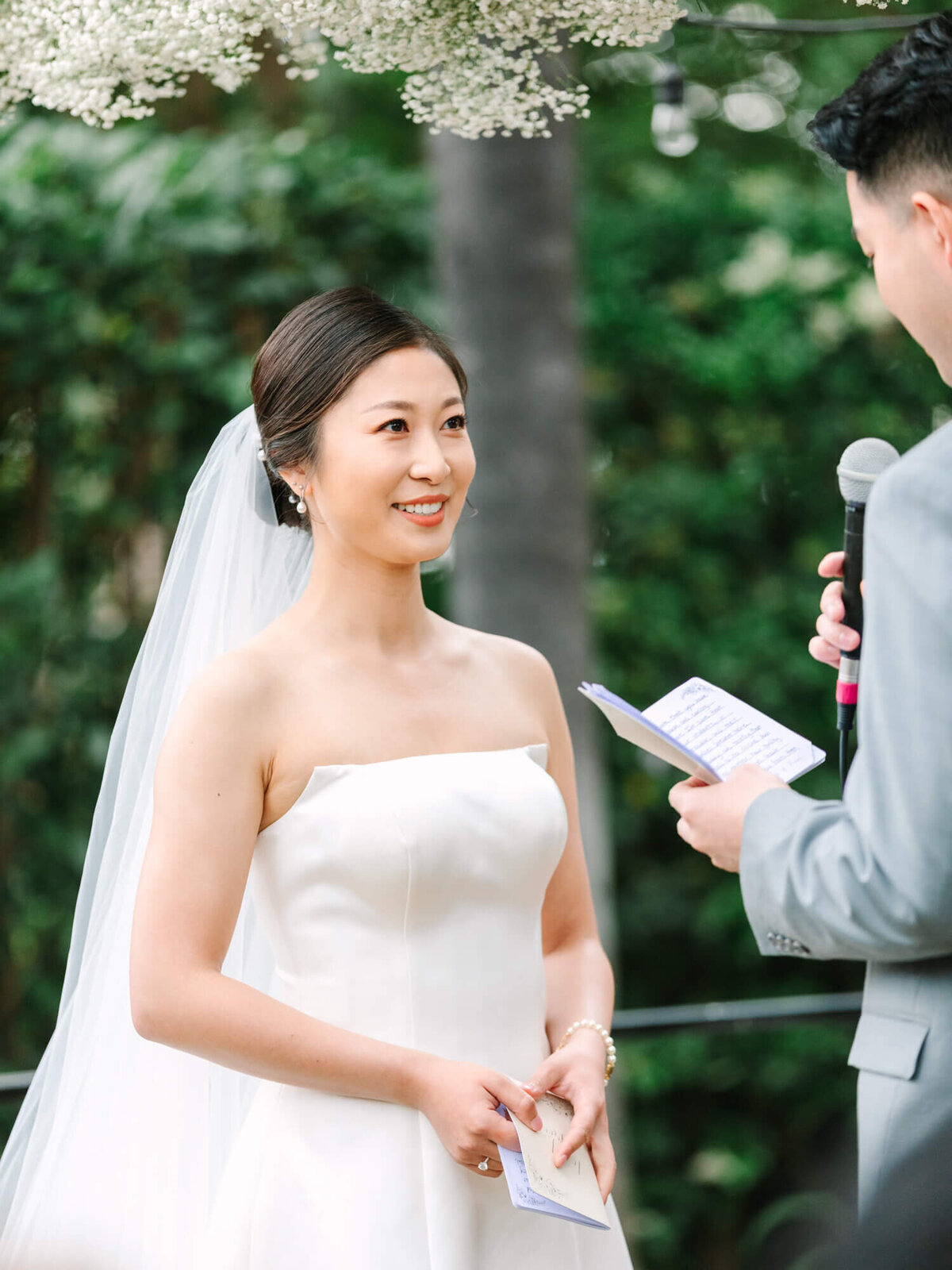 A bride in a white dress and veil smiles tenderly at the groom while he reads vows from a notebook. They stand under a floral arch, surrounded by greenery.