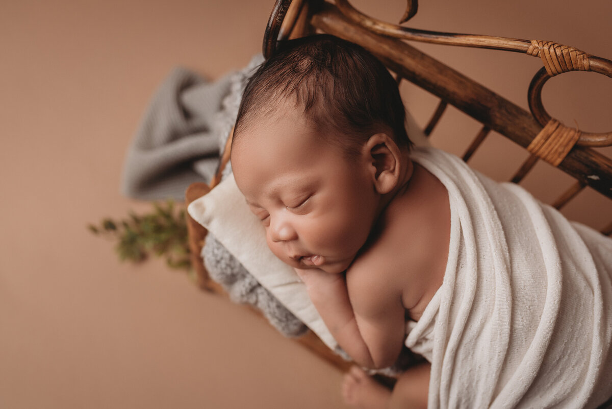 Newborn boy asleep resting on tummy with hand under cheek propped on little bench on tan backdrop
