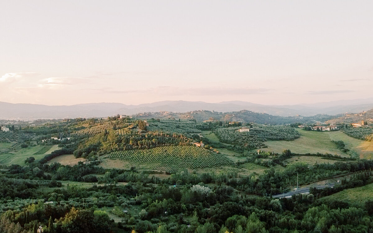 Golden hour view of rolling green hills in the Italian countryside, with layers of vineyards and olive groves stretching toward the distant mountains.