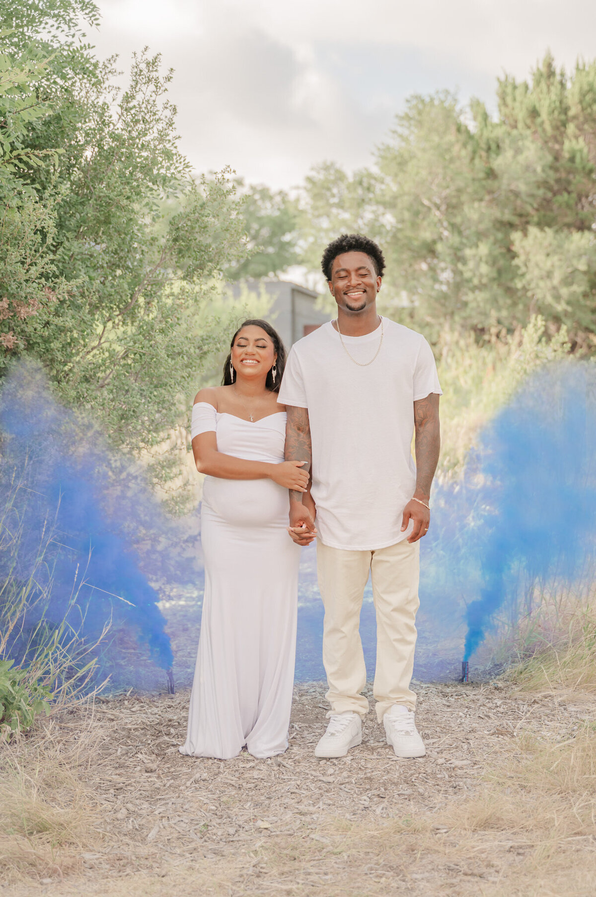 Couple in white stands with eyes closed as blue gender reveal smoke goes off behind them during maternity photos.