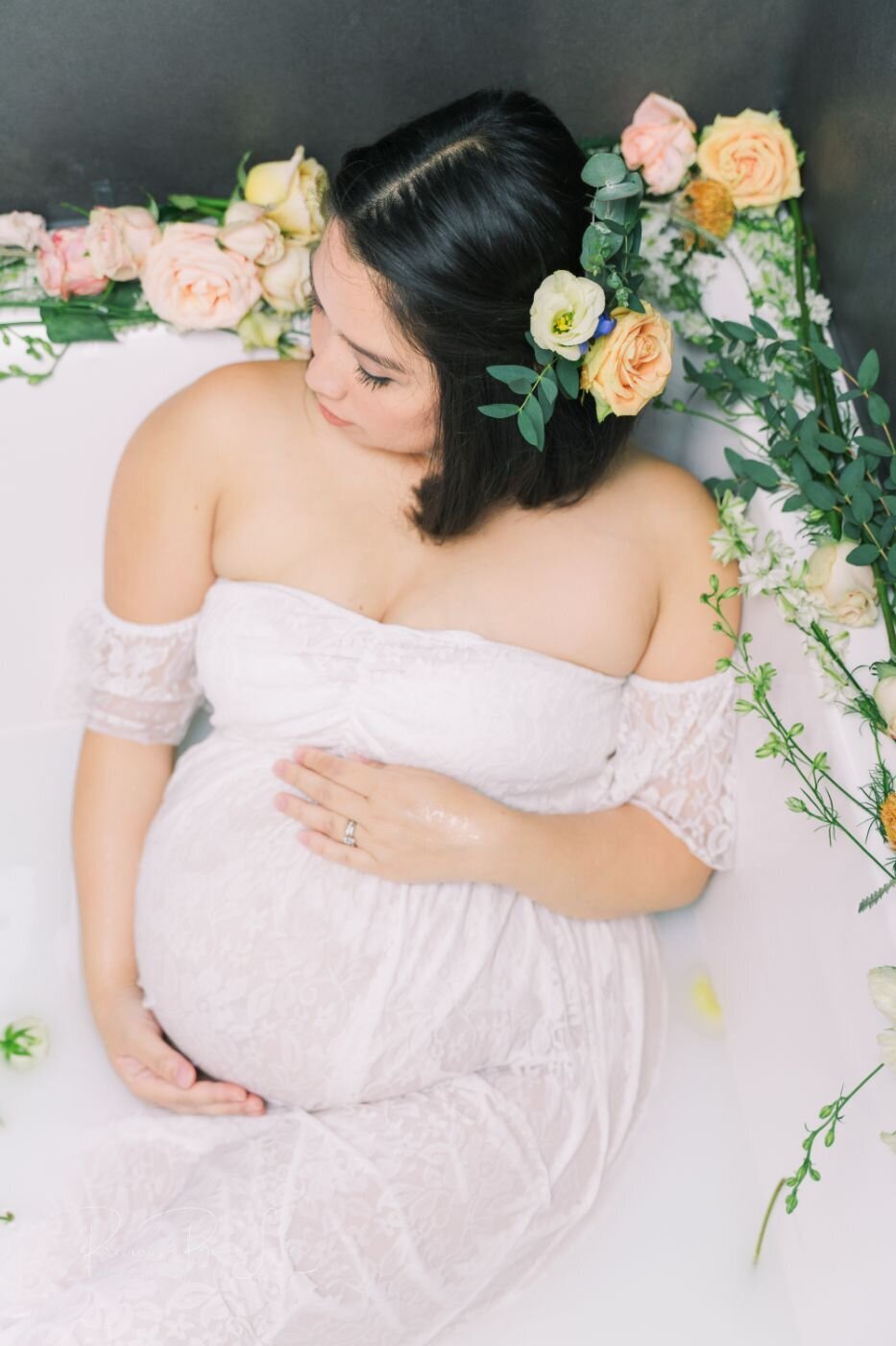 Mother-to-be relaxing in a milk bath surrounded by pastel roses and greenery, wearing an off-shoulder white lace gown and caressing her belly.