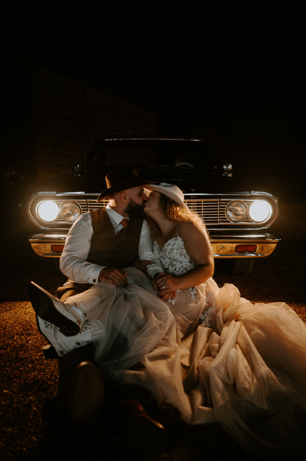 A bride and groom wearing cowboy hats and boots sat inform of an El Camino with its headlights on at night.
