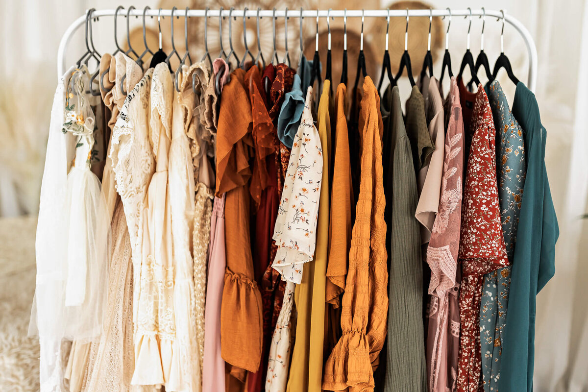 A clothing rack with various dresses in earthy tones, including beige, tan, rust, mustard, olive, and teal. The fabrics are patterned and solid, with textures like lace, floral prints, and ruffles, creating a stylish and diverse collection.