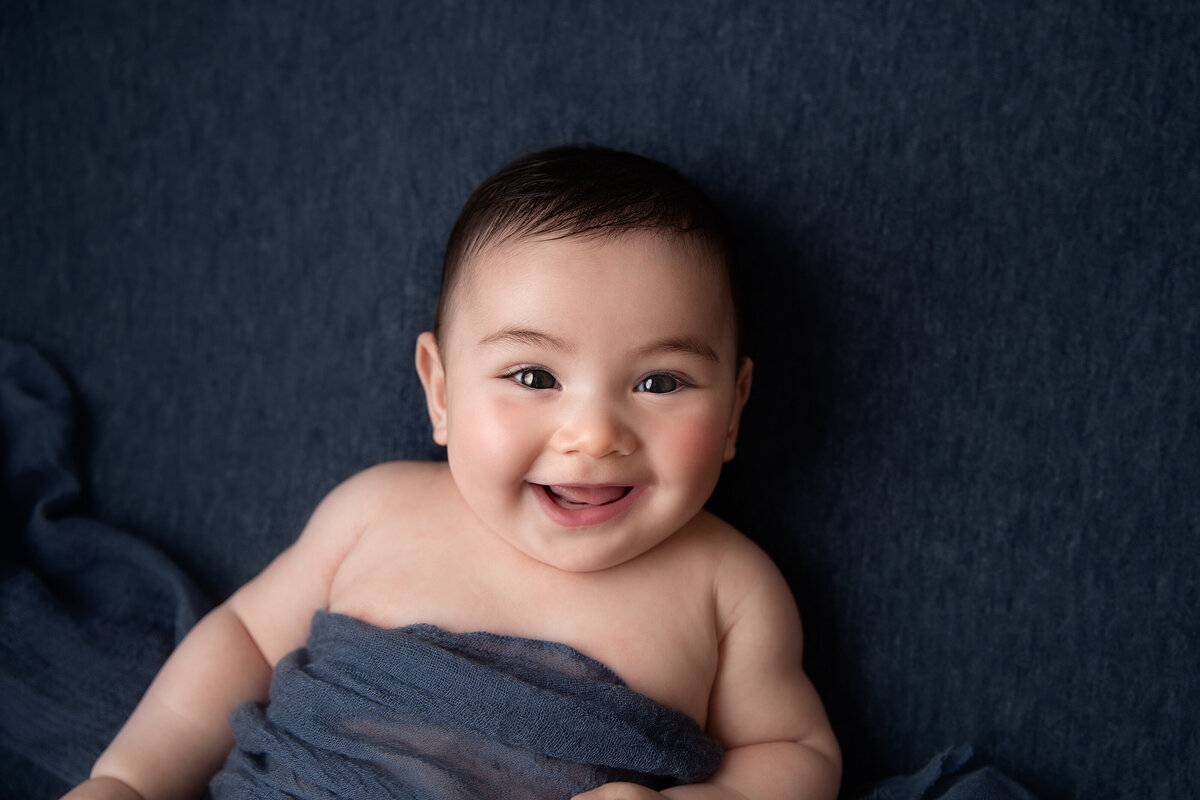 Smiling baby boy lying on a navy blue fabric backdrop with a matching cheesecloth wrap over him.