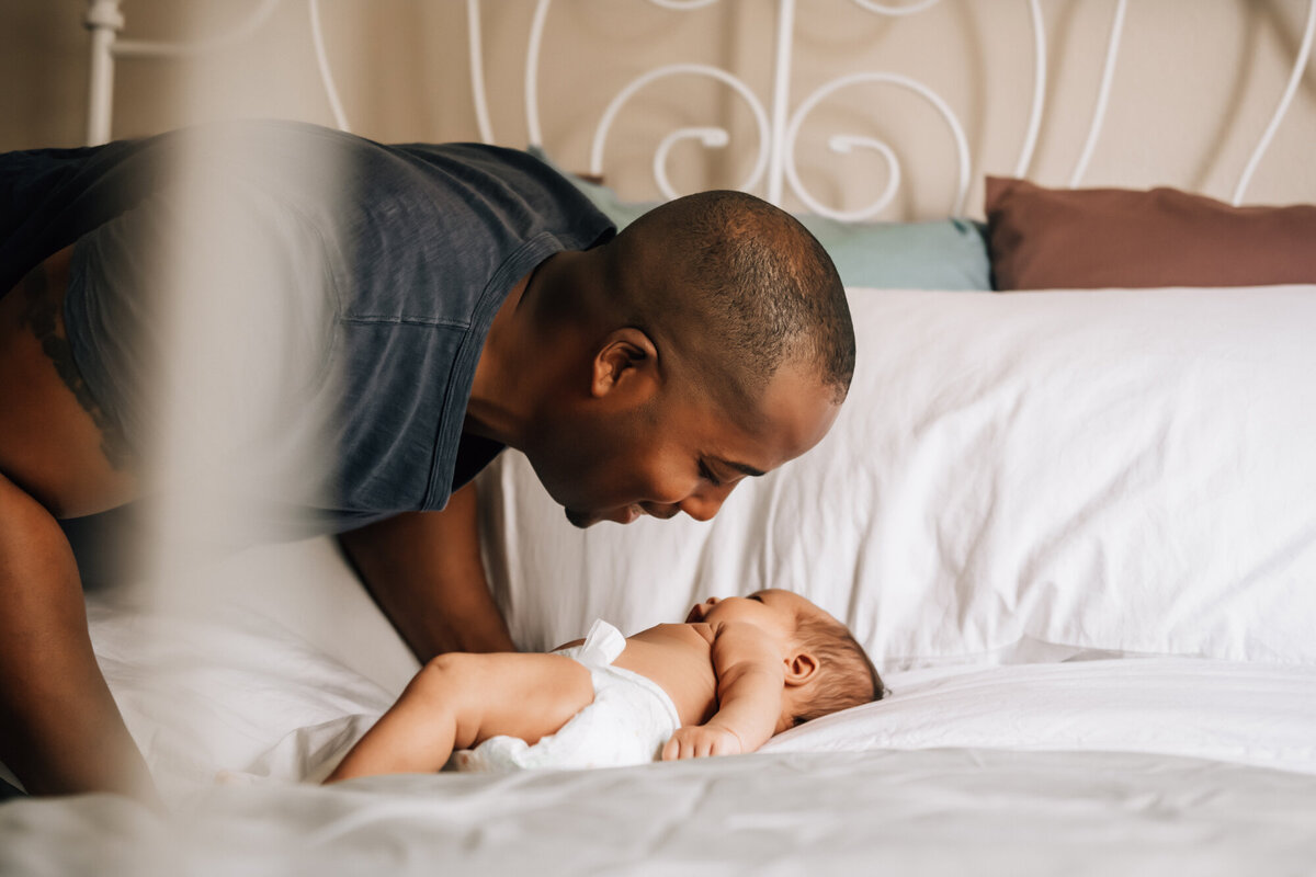 Newborn Photography, Dad leaning over his baby girl, who is laying on the bed, and smiling at her.