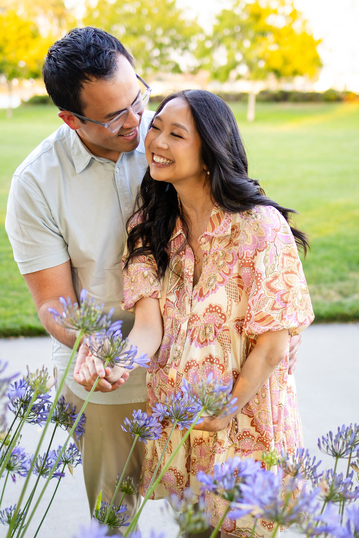 Expecting couple laughs while standing in the garden, gently holding hands near soft purple blooms — Bay Area Maternity Portfolio - Ellobelle Photography.