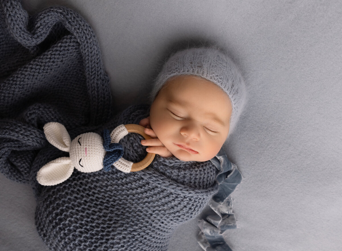 Baby boy in blue knit swaddle is holding a tiny bunny rattle with his fingers peeking out of the swaddle. Baby is sleeping and laying atop of a blue stretch fabric. Captured by top Brooklyn newborn photographer Chaya Bornstein