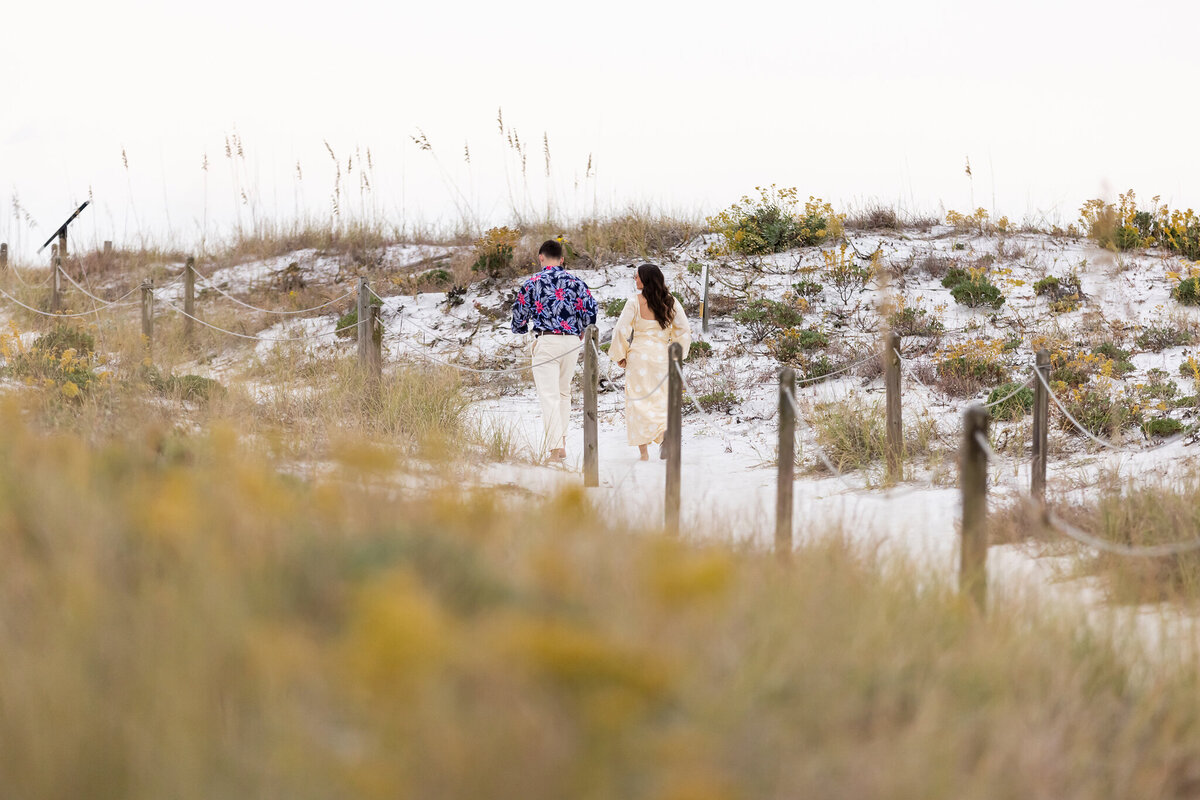 man walking his girlfriend down the path to the beach in Grayton before he surprises her with a proposal while family watches.