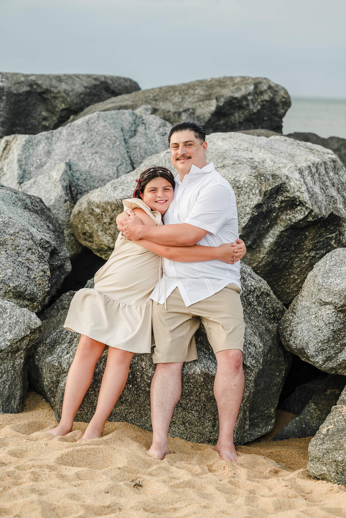 A father, wearing khakis and a white shirt, sits on some beach rocks with his daughter. She is dressed in an off-white dress. They are hugging each other.