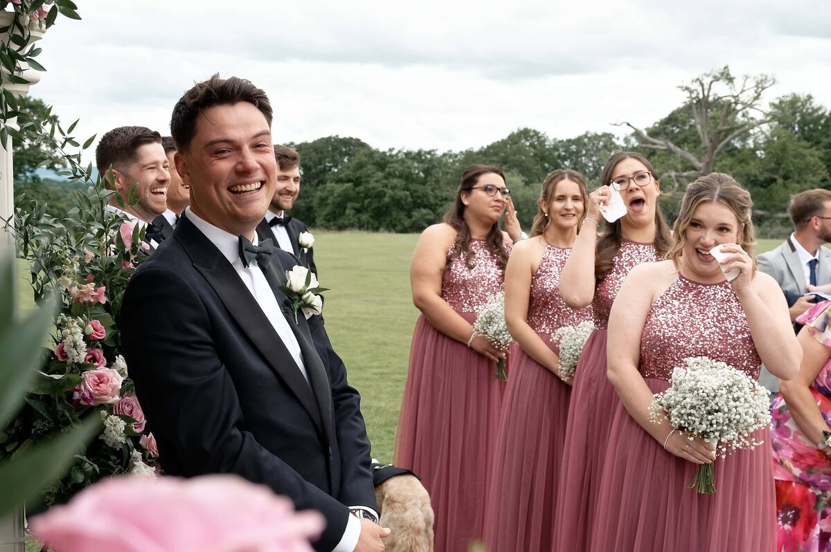 Groom laughing and bridesmaids crying as they wait for the bride to walk down the aisle at South Lodge wedding venue