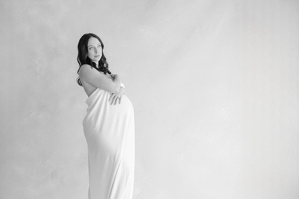 Black and white image of a pregnant woman wrapped in a white blanket looking off in the distance for her Indianapolis boudoir pictures. 