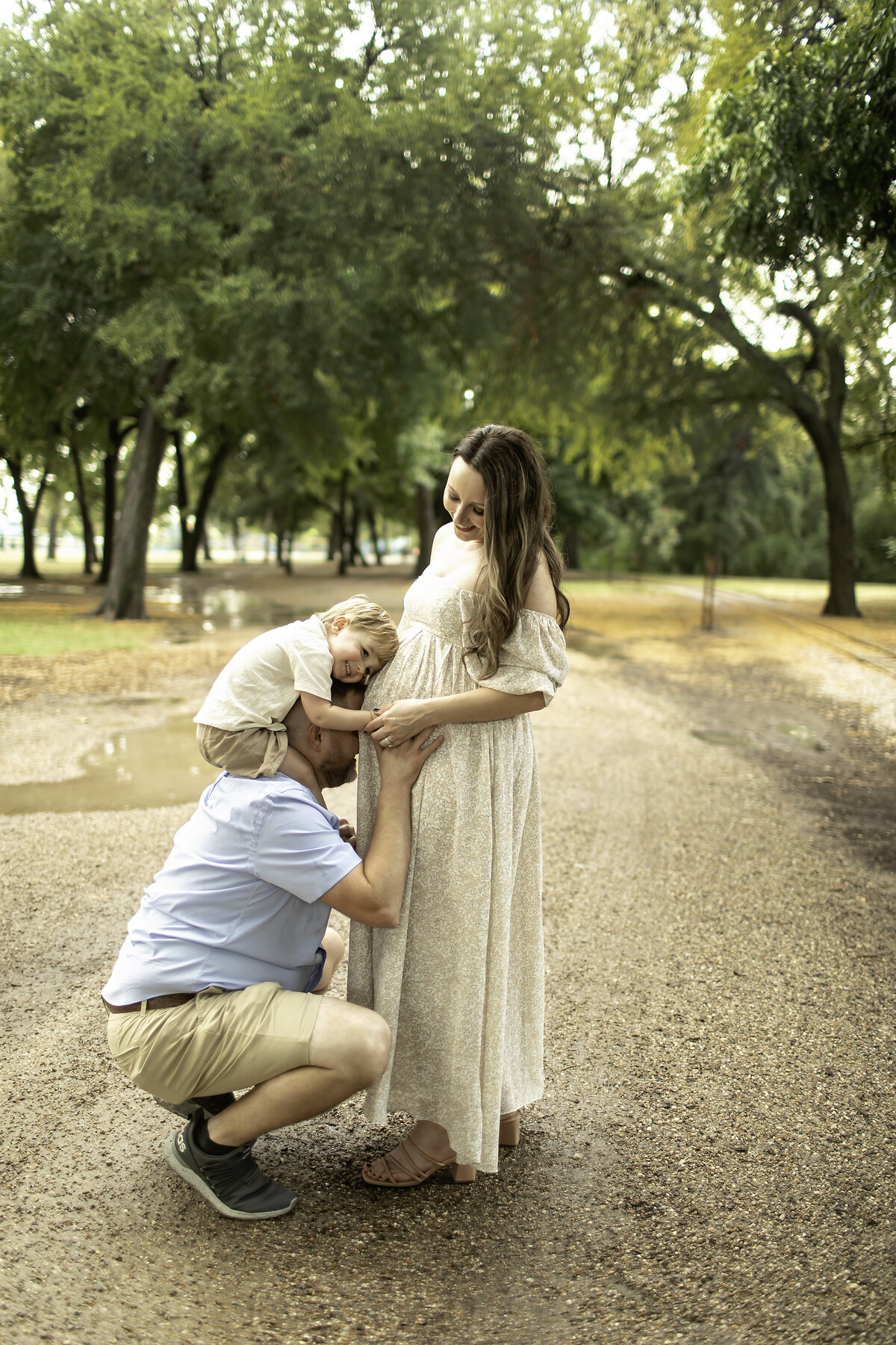 A heartwarming maternity photo at Trinity Park, with dad kissing mom’s belly while big brother sits happily on dad’s shoulders, all sharing a joyful moment together, beautifully captured by Chunky Monkey Photography.