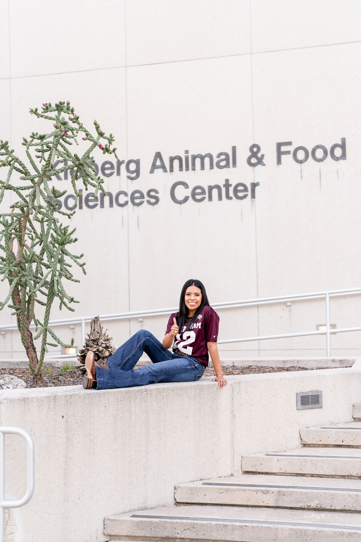 Texas A&M senior girl sitting on ledge at Kleberg wearing jeans and Aggie maroon jersey and giving a thumbs up