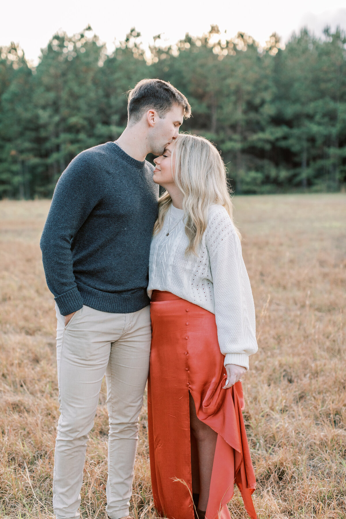 A couple kisses in a field surrounded by pine trees.