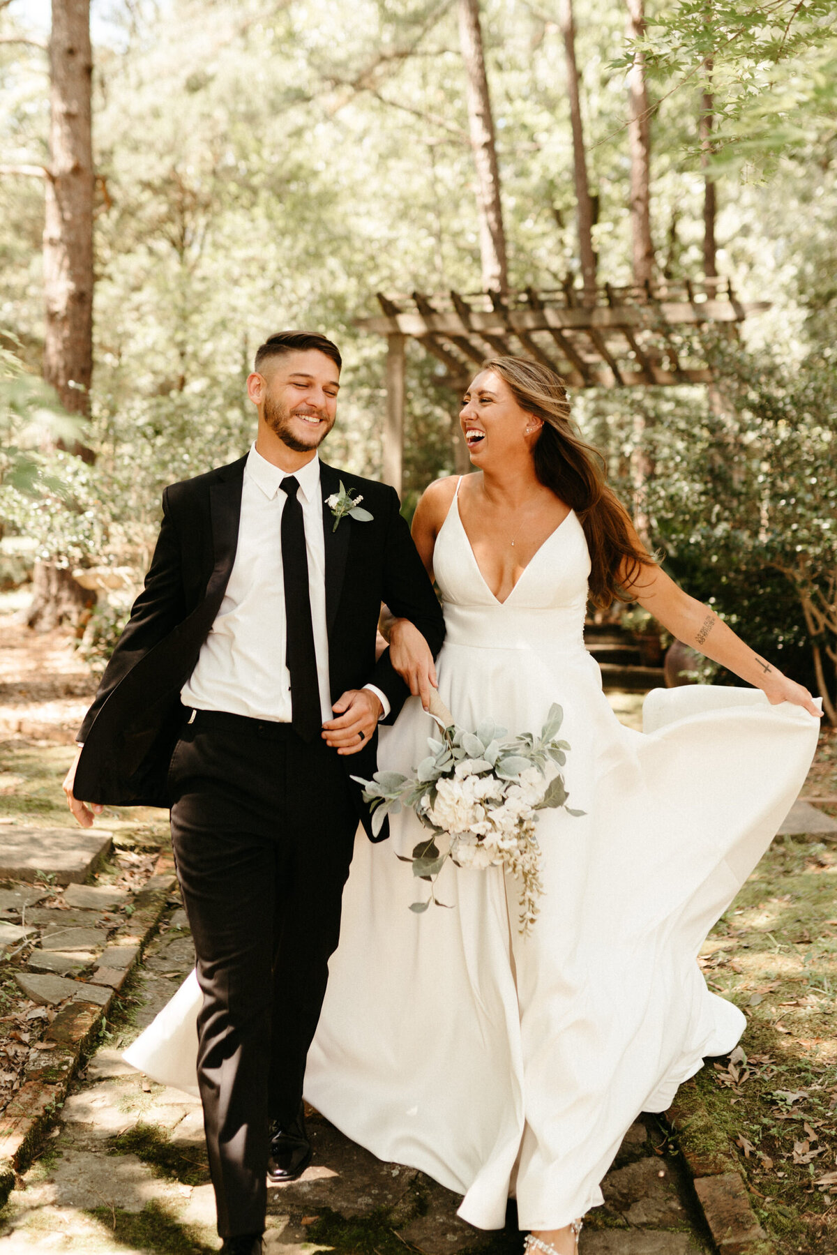 Couple in classic wedding attire laughing and running with trees and a trellis in the background