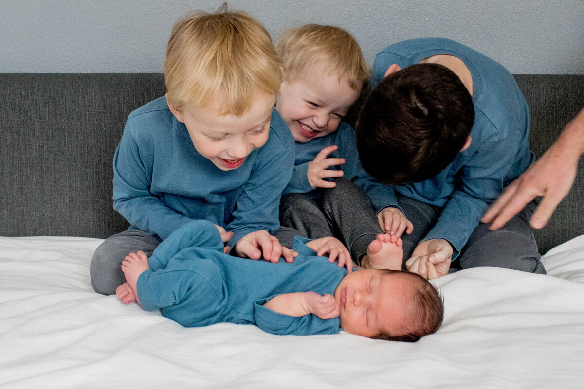 3 young boys in blue tops giggle at their sleeping newborn baby brother