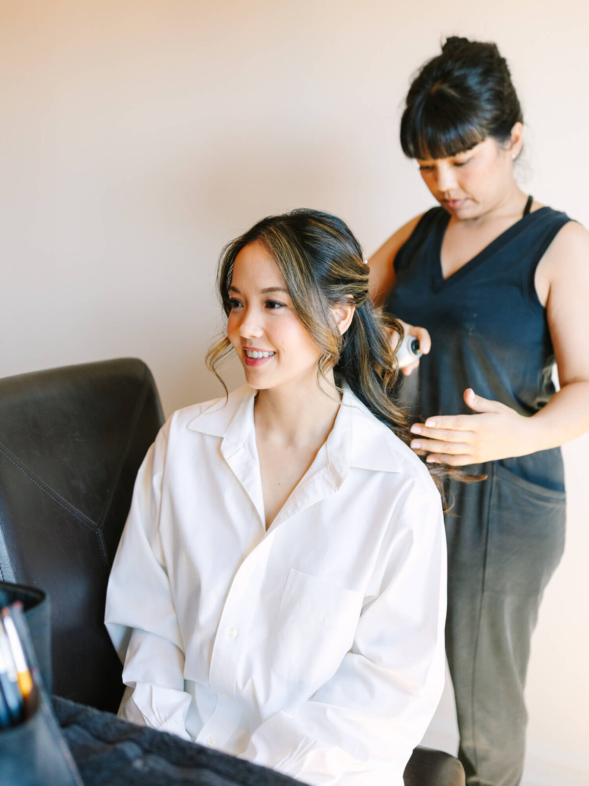 A smiling bride in a white shirt sits on a chair while a hairstylist curls her hair. The setting is calm and joyful.