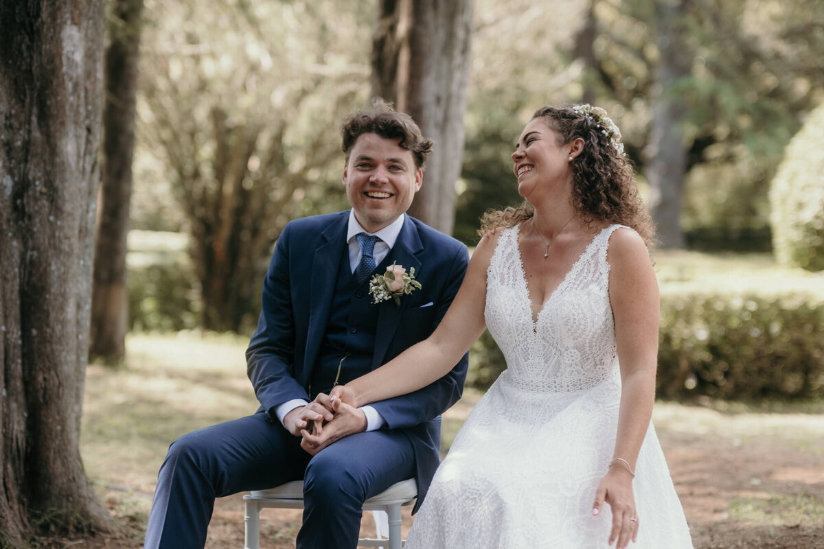The wedding couple sits during their wedding ceremony laughing heartly and holding hands.
