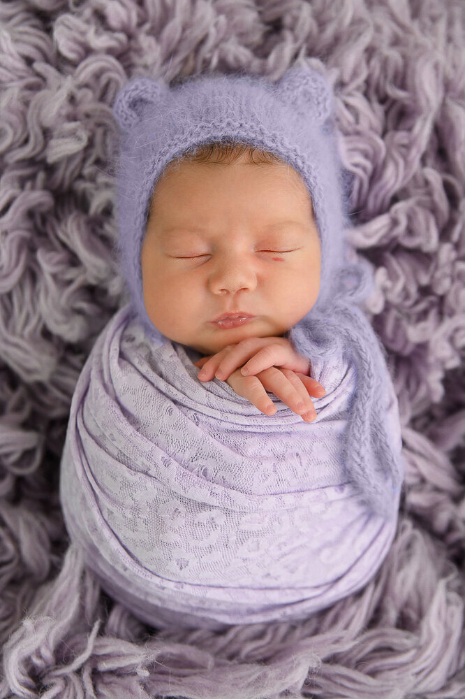 newborn girl wrapped in purple wearing a purple bear hat laying on a purple rug.