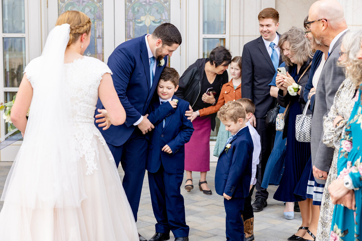 Groom in a navy blue suit hugs his two young sons in matching suits as the bride and family members look on with joy.