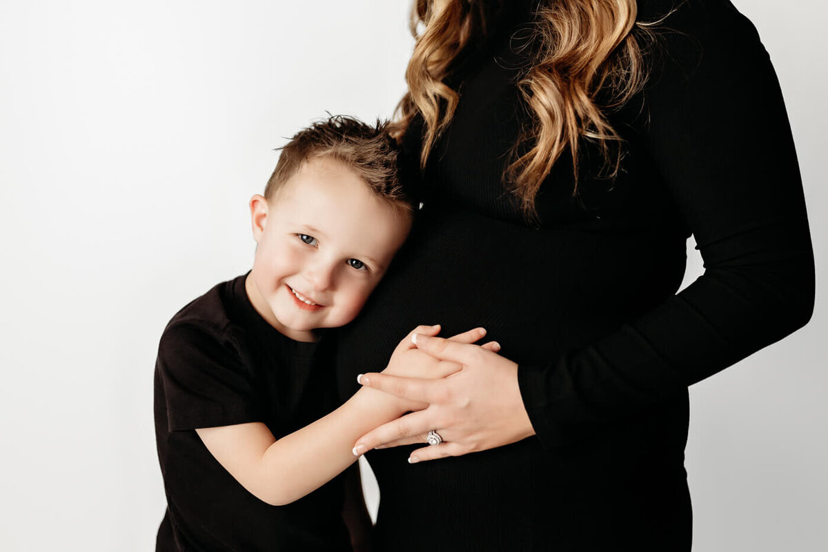 Toddler boy hugging on mom's baby bump in studio against white backdrop
