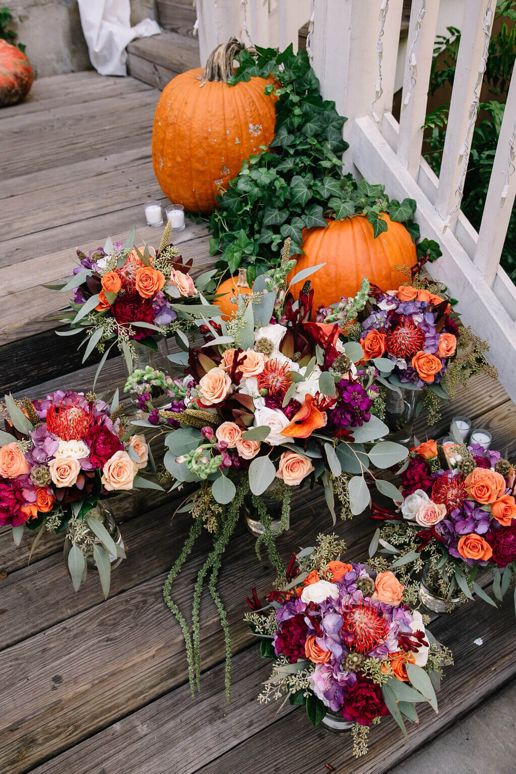Several vibrant floral bouquets featuring roses, eucalyptus, and other colorful flowers are arranged on wooden steps with pumpkins and vines.
