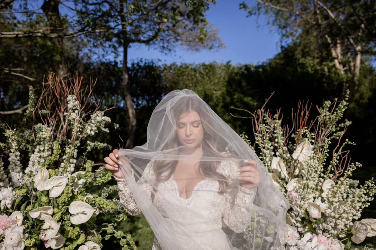 Bride poses with her veil draped over her face in a serene editorial portrait surrounded by sculptural white and blush floral arrangements at a private estate wedding in the Hamptons.