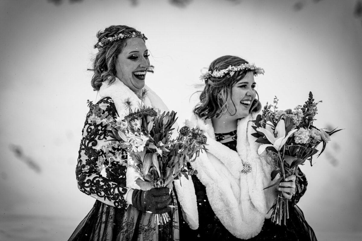 Two brides, both wearing white fur stoles, look out from the top of a mountain in Alaska