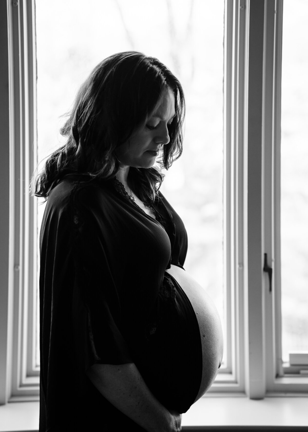 A side profile of a pregnant woman standing by a window, cradling her belly.