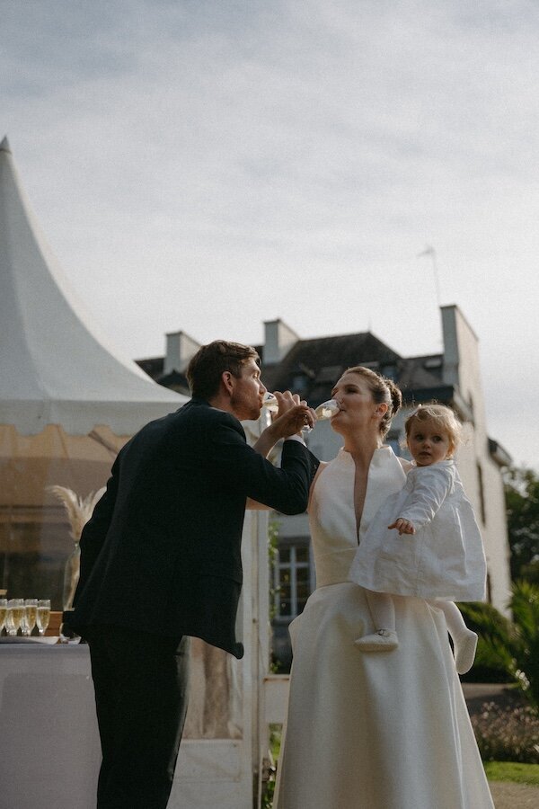 Les mariés trinquent lors du cocktail, la mariée portant leur enfant dans ses bras. Moment festif capturé par Kamescop au Château de Beaussais en Bretagne.