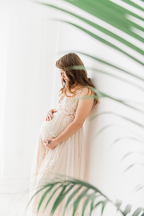 Softly lit studio portrait of a pregnant woman in a lace dress, framed by palm leaves for a tropical, serene vibe.