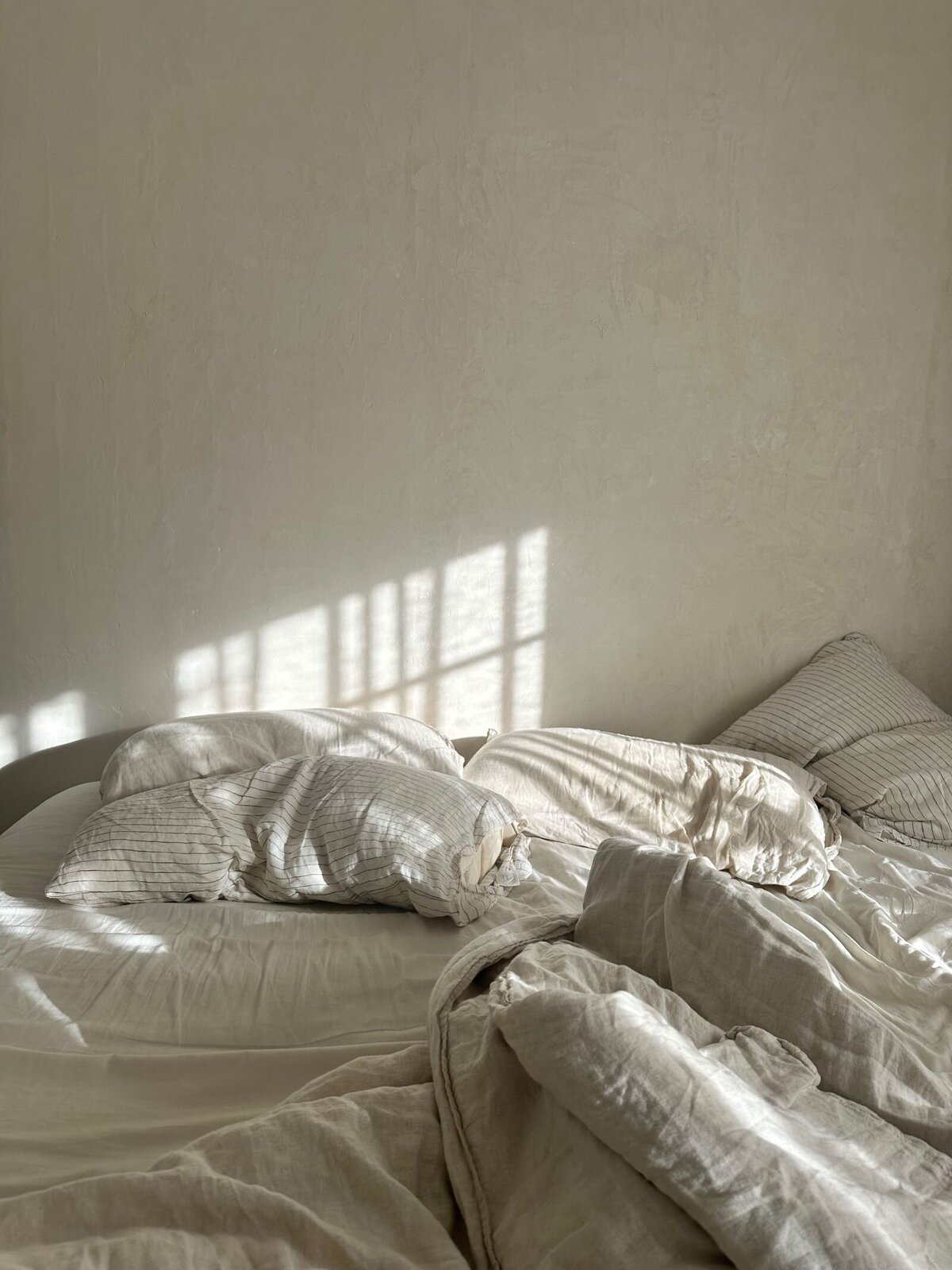 Close-up of an unmade bed with soft neutral linens, set against a warm beige plaster-covered wall, exuding a relaxed, serene atmosphere.