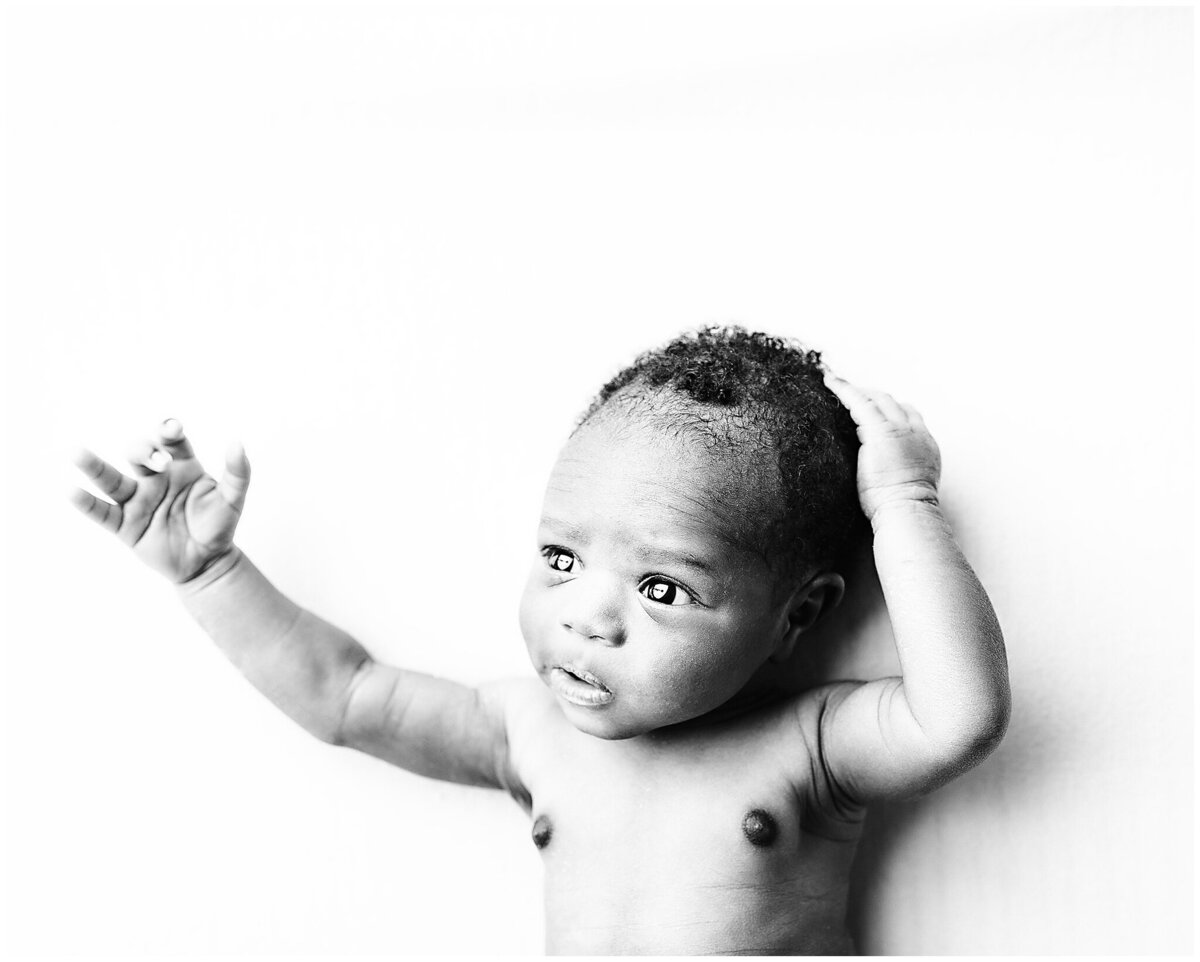 Baby boy laying on his back with one hand on his head and the other arm out stretched looking at the light.