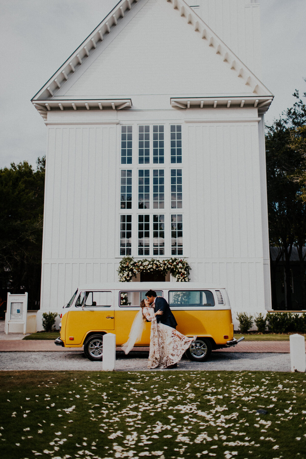 A bride and groom kiss while embracing in front of a yellow vw bus