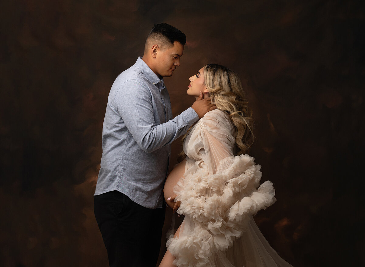 A man in a blue shirt places a hand on his pregnant bride in an open tule maternity gown in a New Orleans Maternity Photography studio