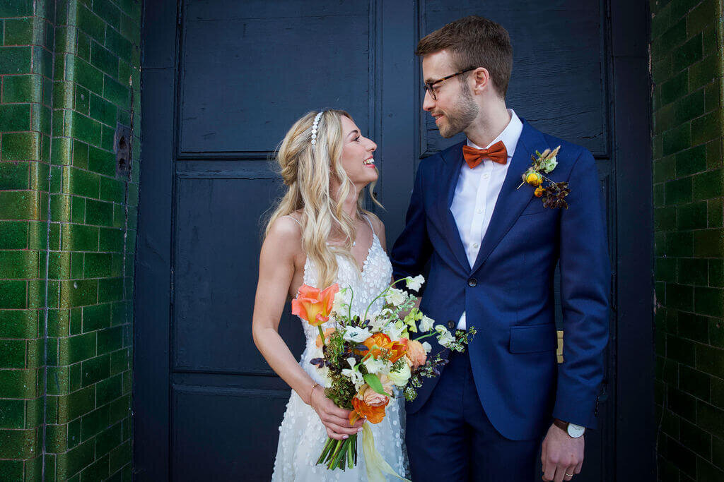 Bride and groom standing in Hackney Road doorway. Bride holding orange bouquet