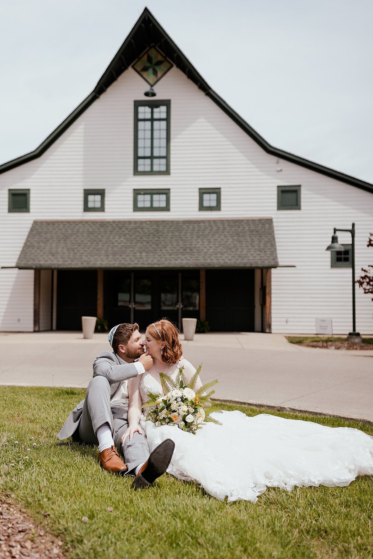 Jewish groom wearing a light grey suit and white kippot kisses a redheaded bride in a flowing white dress. The bride is holding a large bouquet of fern and wild flowers in front of a large white barn with green trim at Loveless Cafe in Nashville, TN
