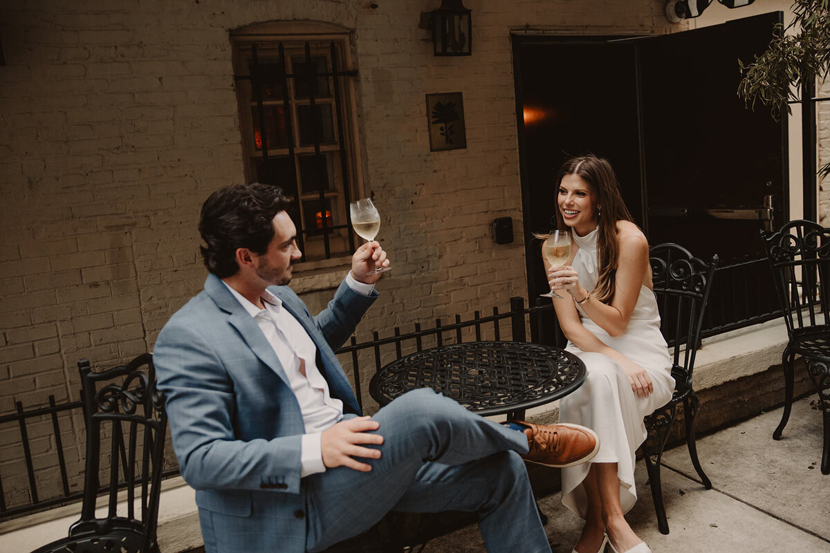 A couple sitting at a small table holding up wine glasses in a toast 
