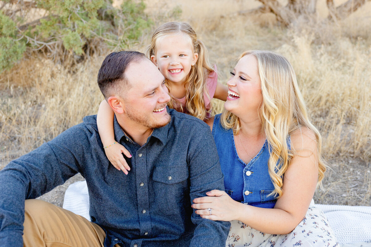 A young girl wraps her arms around her parents while they sit on a blanket, all laughing and enjoying the moment together. The warm, golden light adds to the joyful atmosphere.