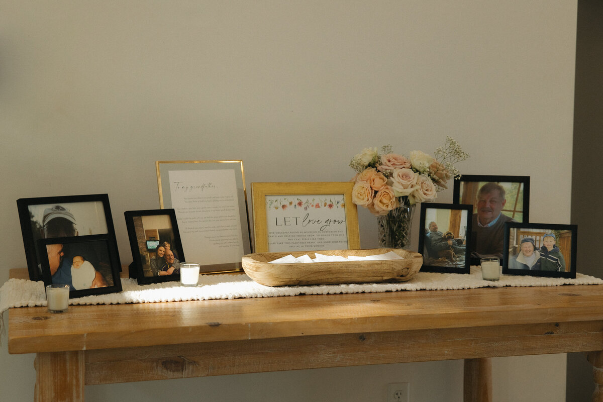 A wooden table set up as a memorial for a wedding reception. There is a bouquet of light pink, light orange and white roses, old photographs of family members and small tea candles. There is a sign that says let love grow where wedding guests can take from a small wooden bowl in front with white packets containing seeds. 