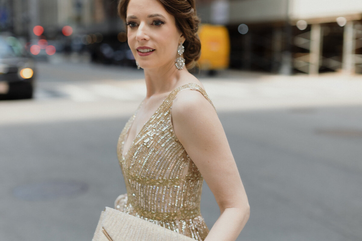 close up shot of woman walking across the street in urban area, wearing a gold gown and carrying a clutch.