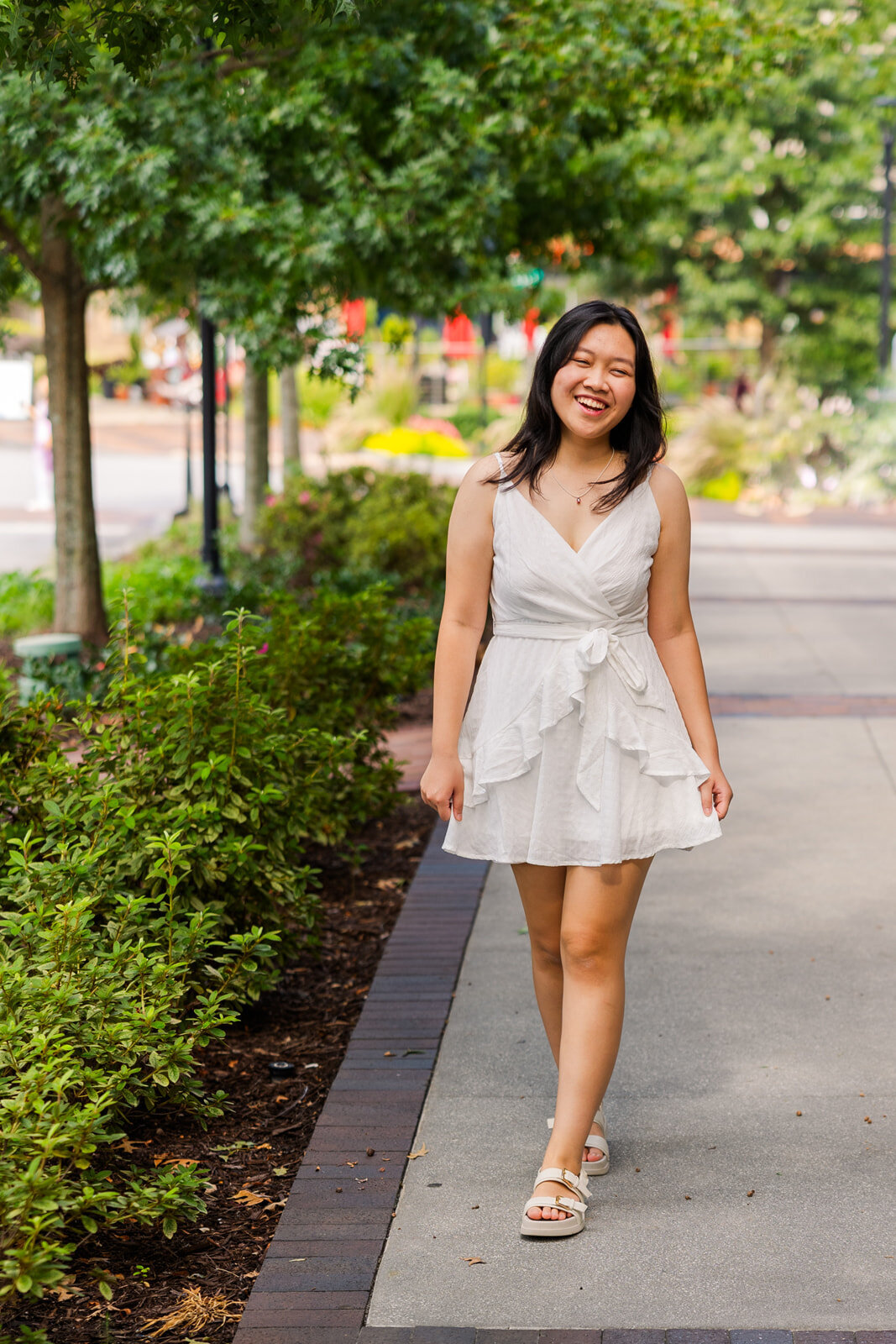 Senior girl wearing a short white dress walking and laughing on a street sidewalk during portrait photo session