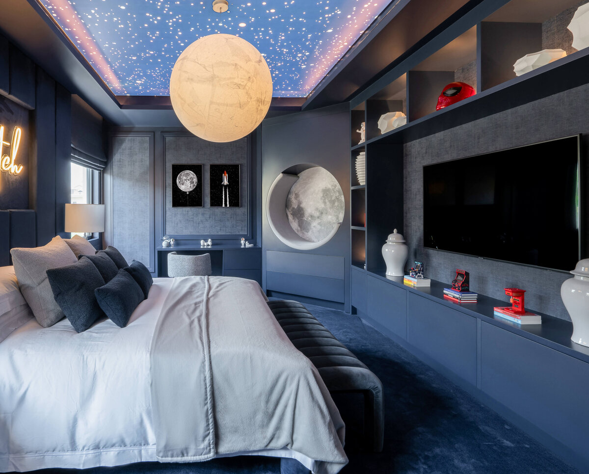 Boy's room with dark blue outer space theme. The ceiling is illuminated to look like the night sky, and the main light fixture looks like the moon. The bed to the left is adorned with blue bedding. There is a television on the wall across from the bed. In the background, and built-in chair is cut into the wall and painted to look like the moon.