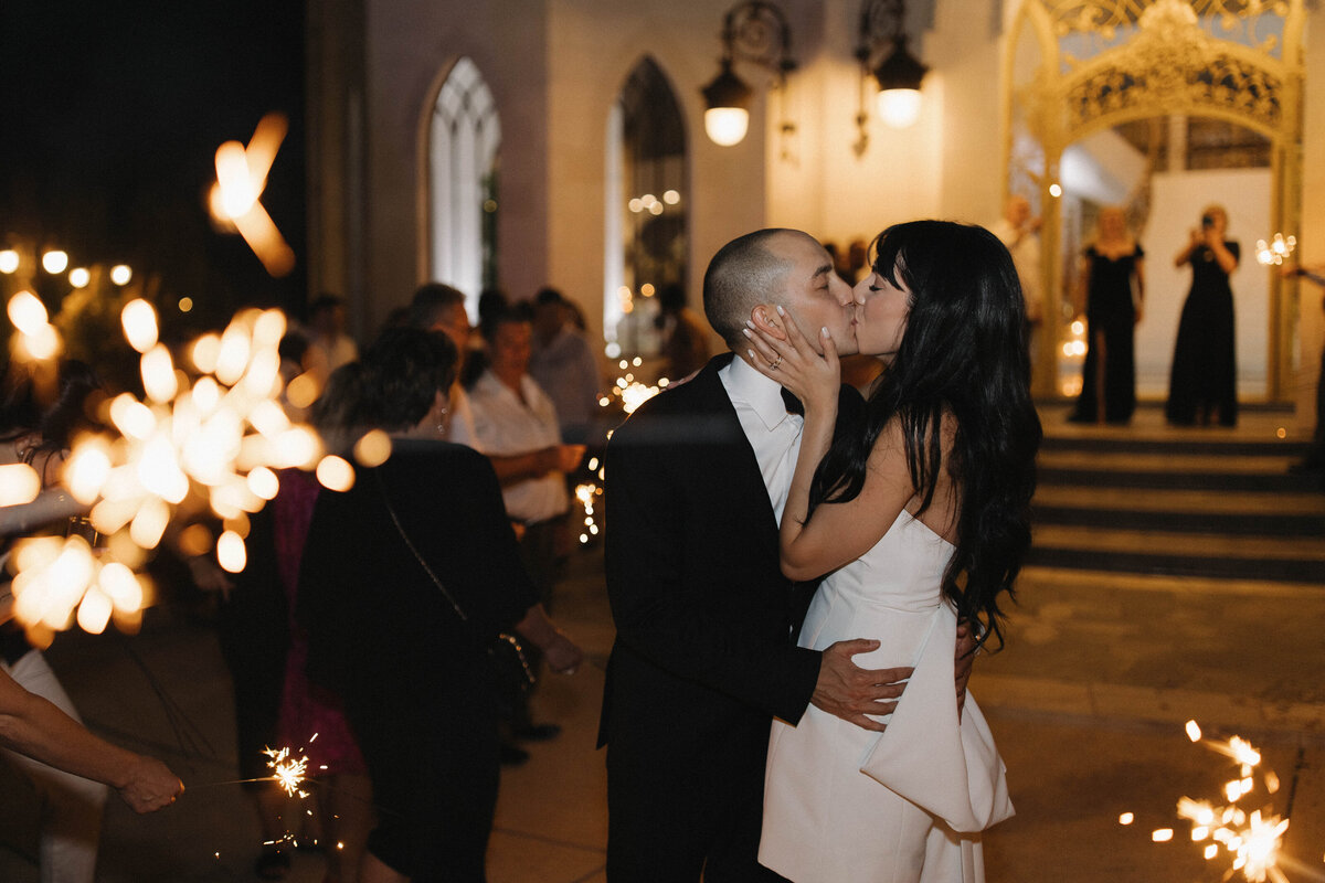 A newlywed couple shares a kiss during their sparkler exit, surrounded by guests holding glowing sparklers. A cinematic and documentary-style wedding celebration photo.