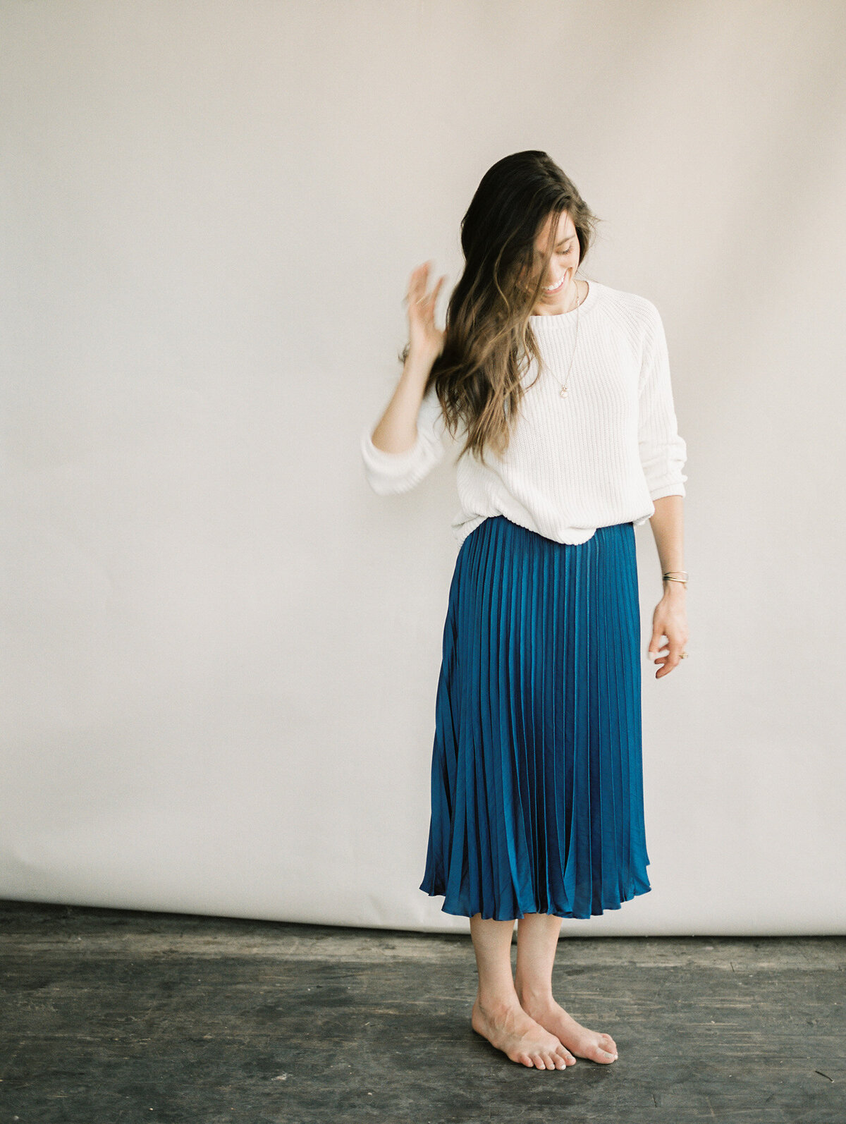 barefoot woman in cream sweater, blue pleated skirt, soft beige backdrop, studio photography