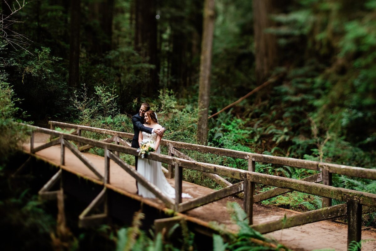 A bride and groom embrace on a charming wooden bridge in Redwood National Park.